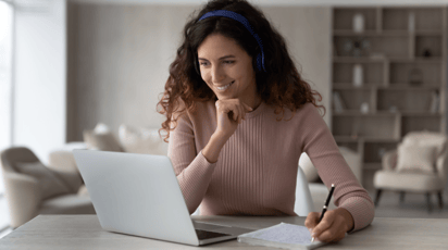 Young woman at a desk with her laptop writing notes in a notepad 